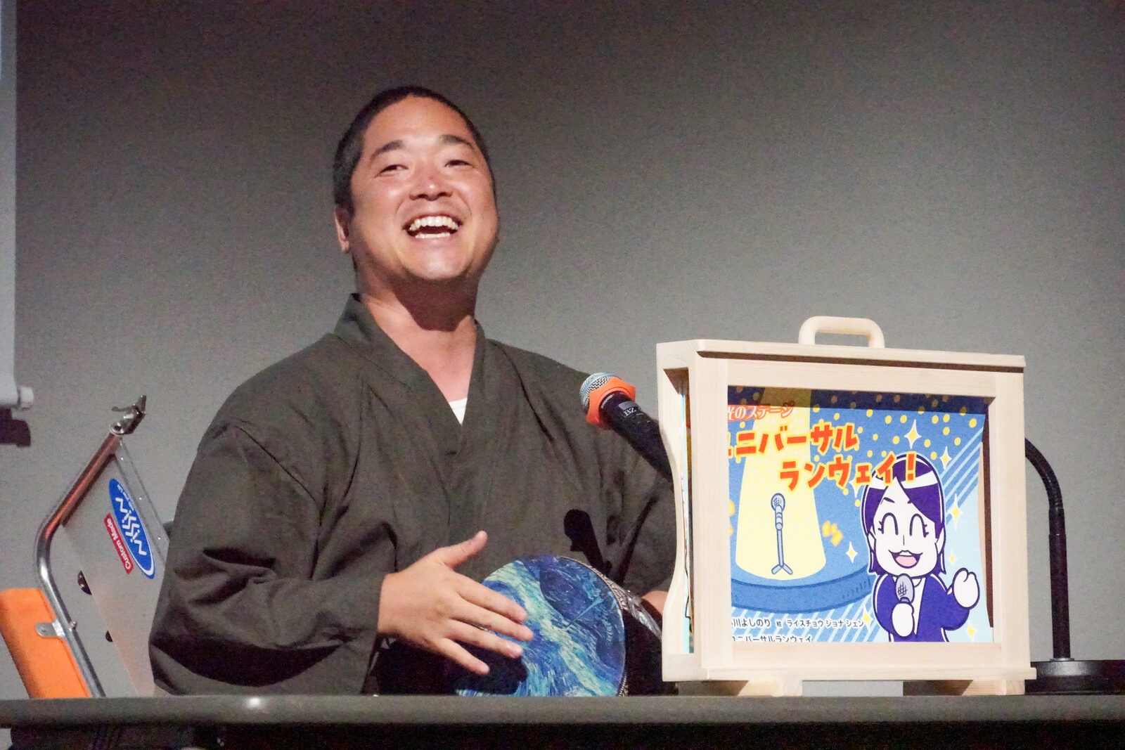 和装の小川よしのりさんが、舞台上で紙芝居の枠を横に置き、身振りを交えて語っている。