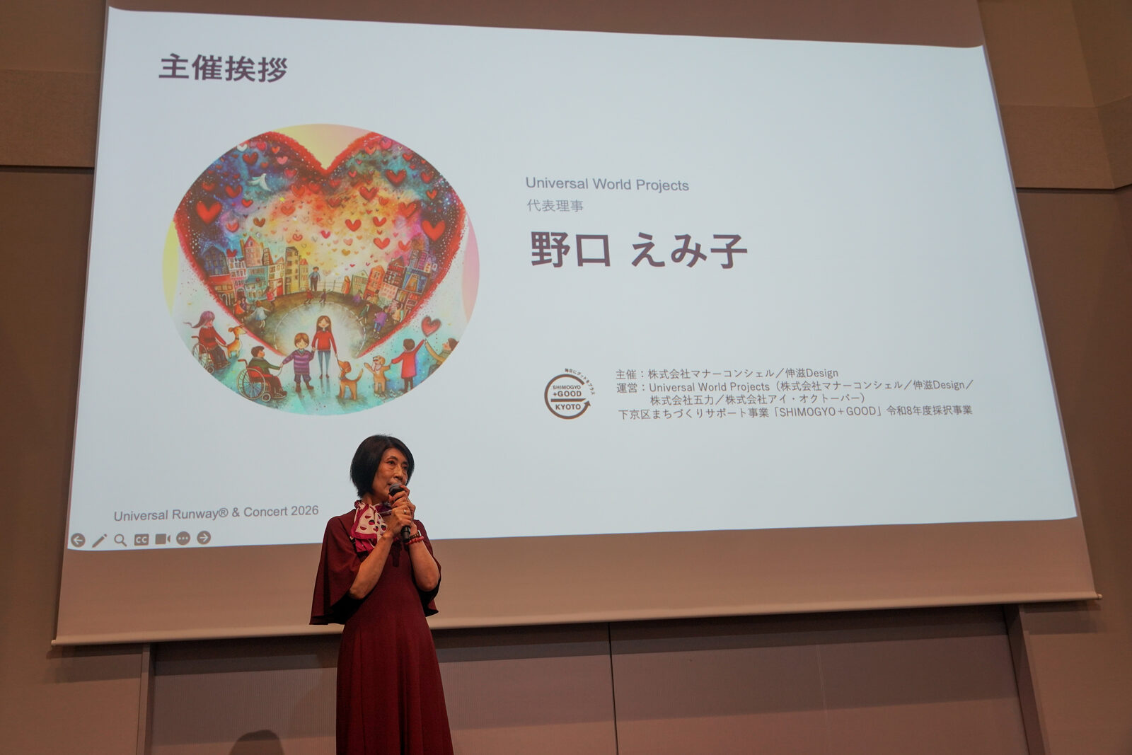 赤いワンピースの野口えみ子さんがマイクを持って話している。背面の大型スクリーンには「主催挨拶 野口 えみ子」のスライド。