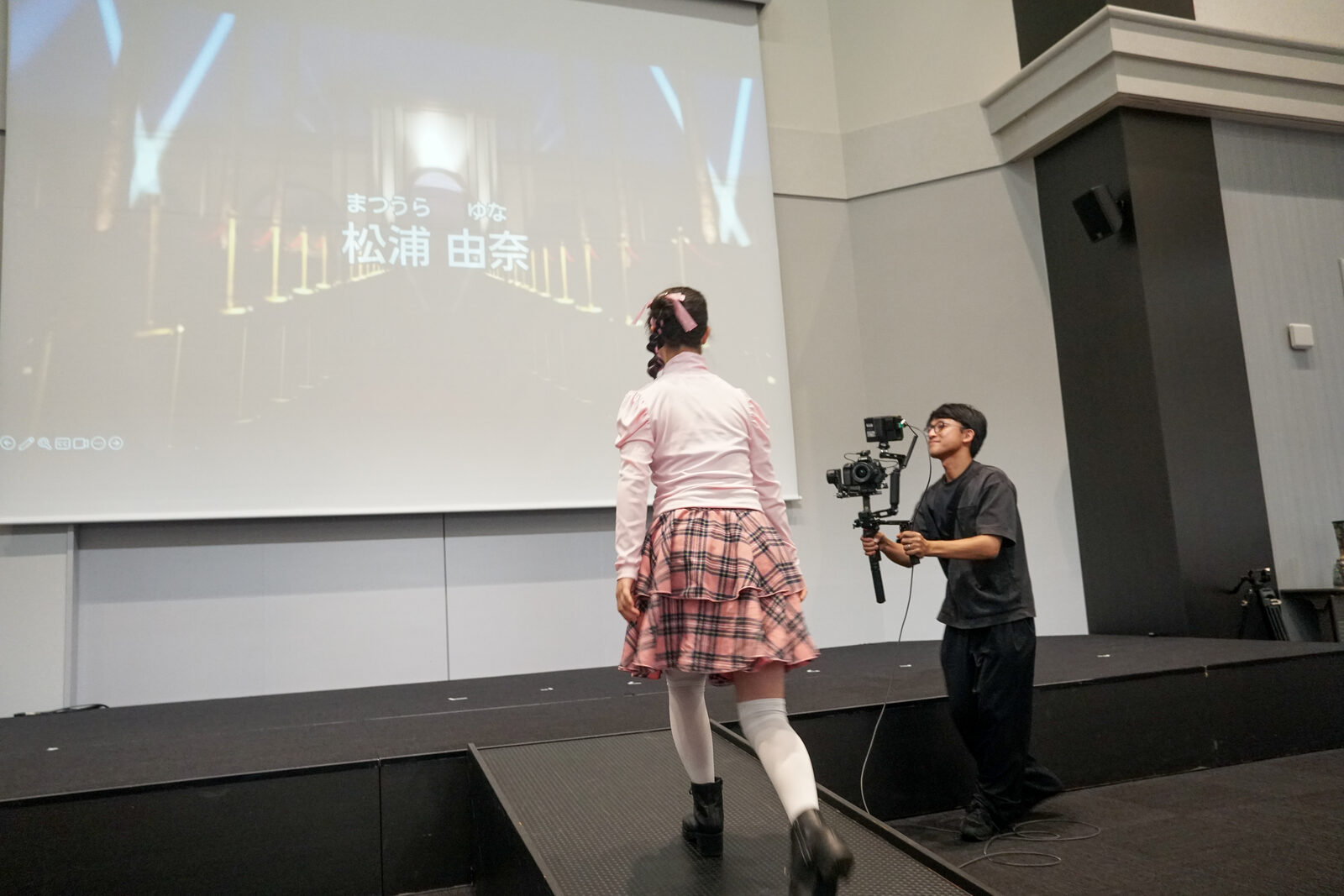 ピンクの衣装の松浦由奈さんがランウェイを歩いている。スクリーンには名前が投影されている。