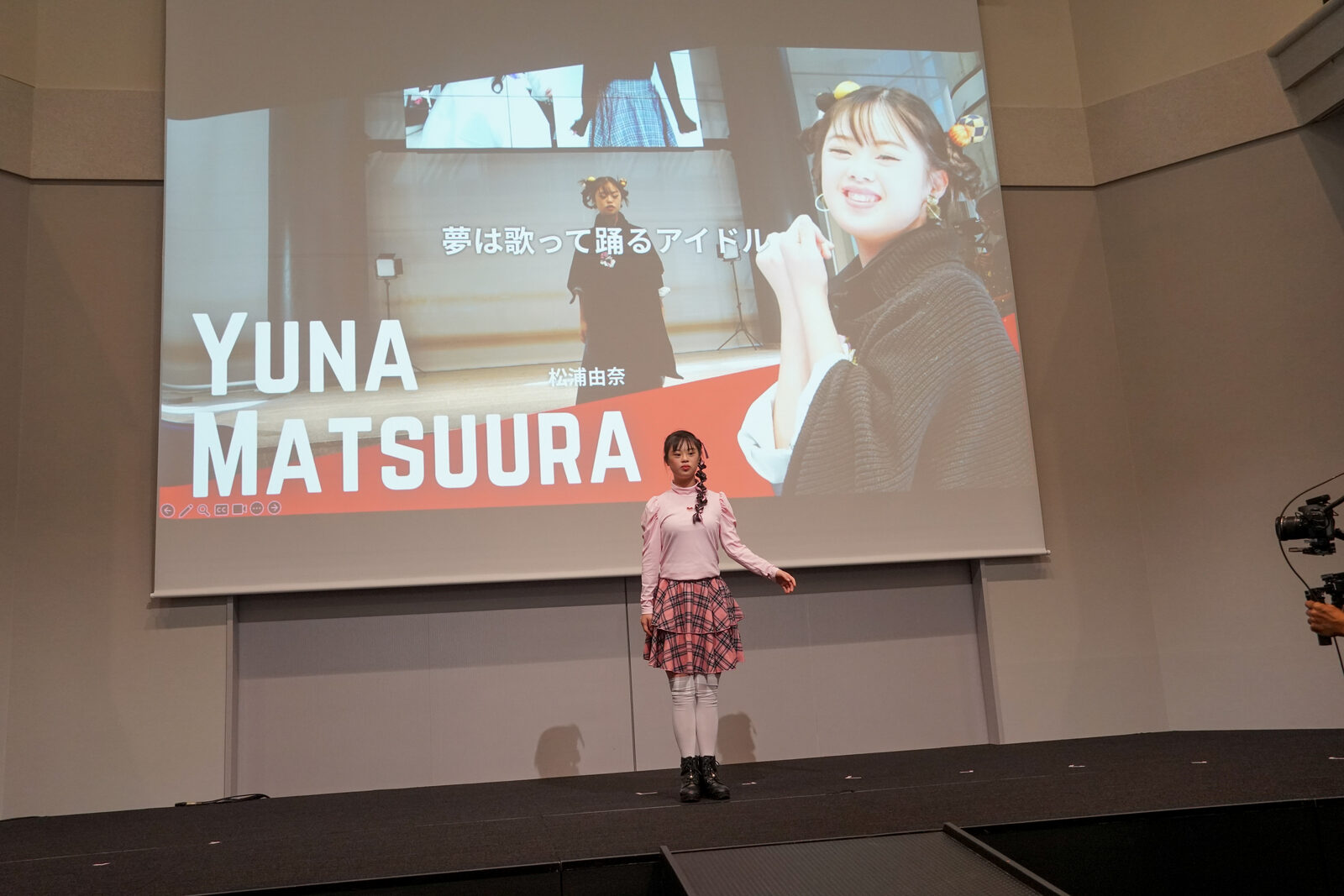 スクリーンに YUNA MATSUURA の紹介映像が大きく映り、その前に松浦さんが立っている。