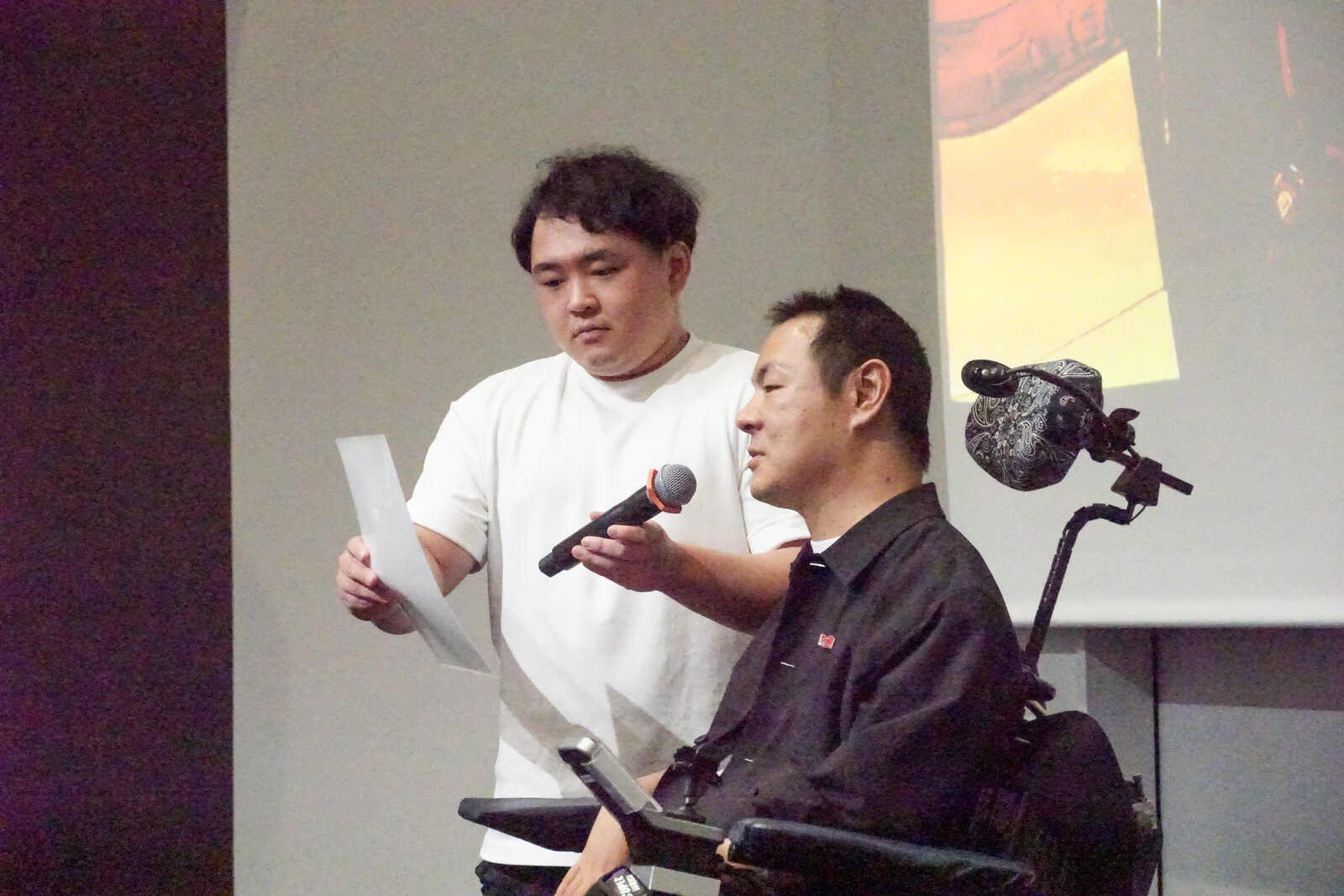 電動車椅子の伊藤靖幸さんが、サポーターの男性とマイクを分け合って話している。