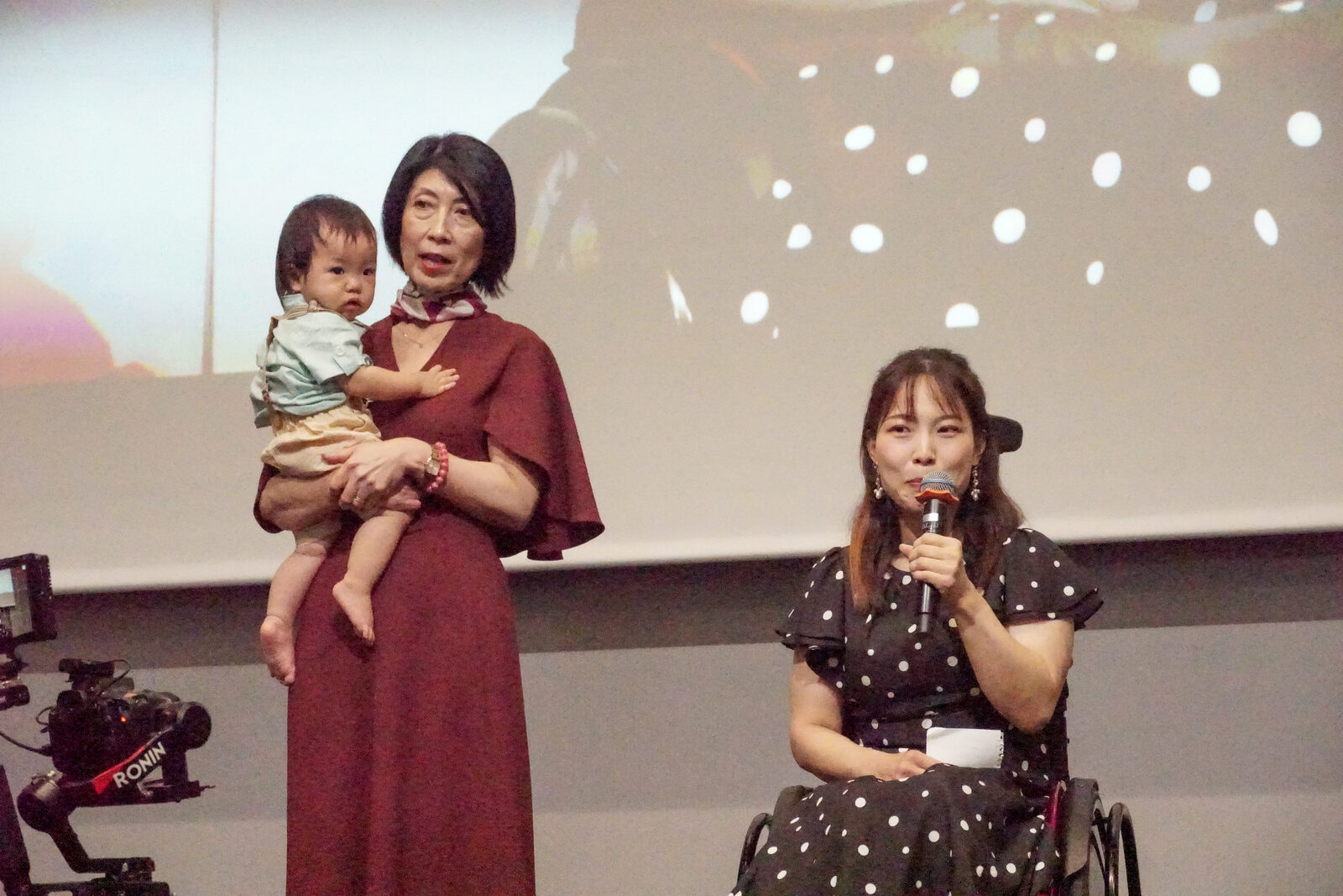 野口えみ子さんが赤ちゃんを抱き、その隣で愛澤咲月さんがマイクを持って話している。