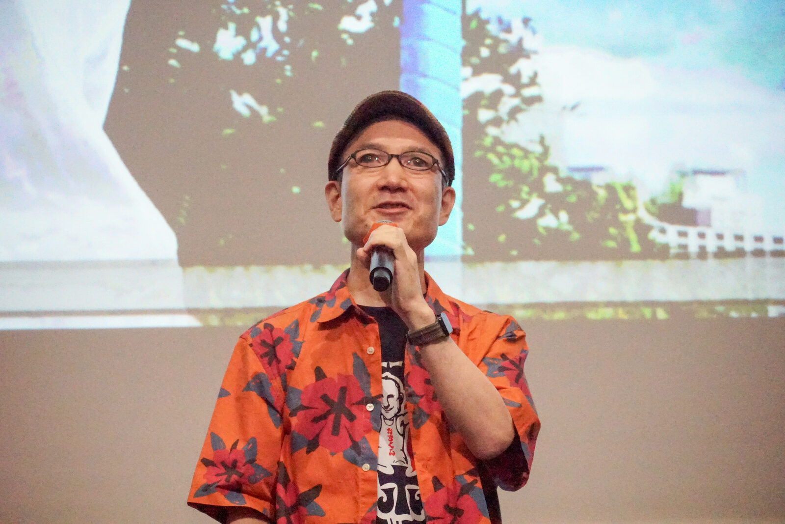 オレンジ色のアロハシャツの野元和明さんが、マイクを持って笑顔で話している。