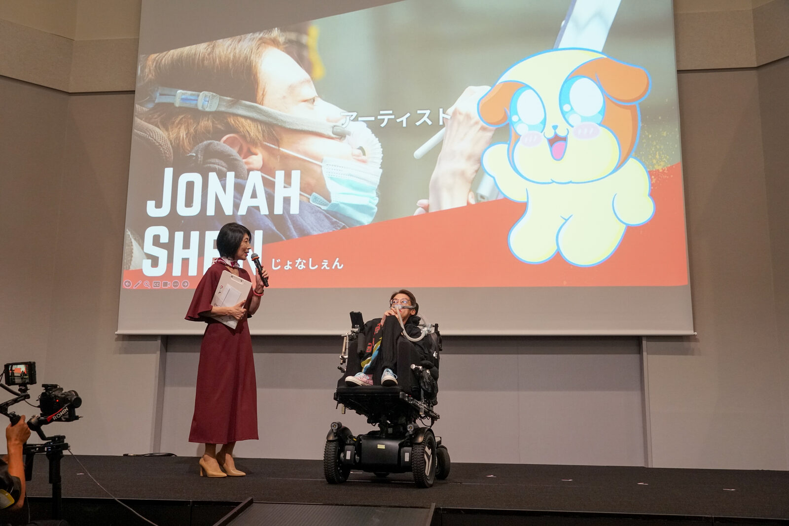 人工呼吸器をつけたジョナ・シェンさんが電動車椅子で登場し、司会と向かい合っている。スクリーンには JONAH SHEN のタイトル。