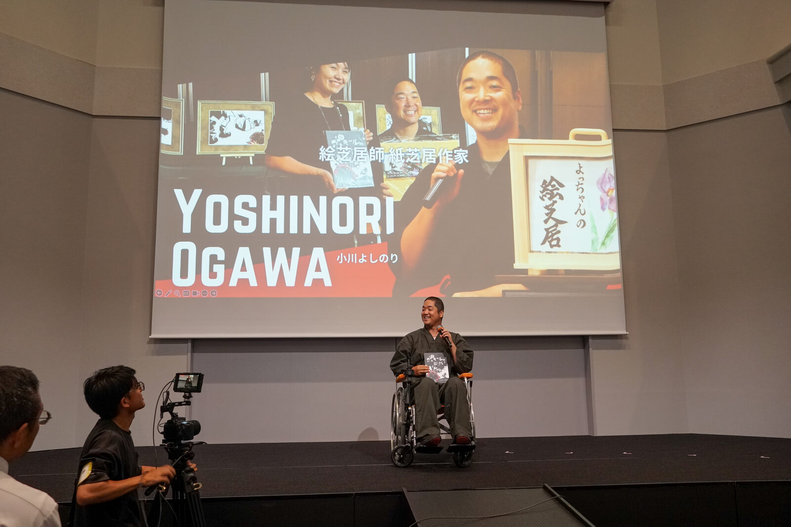 和装の小川よしのりさんが車椅子で舞台に上がり、スクリーンには YOSHINORI OGAWA のタイトルが映る。