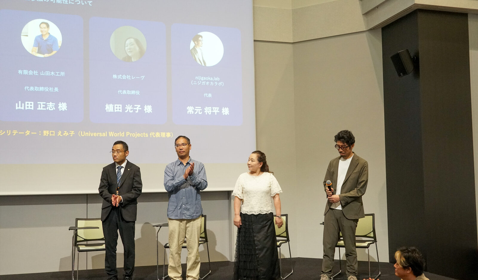 舞台上に4名の登壇者が並び、背面スクリーンには登壇者の顔写真と所属が並んだスライドが映っている。