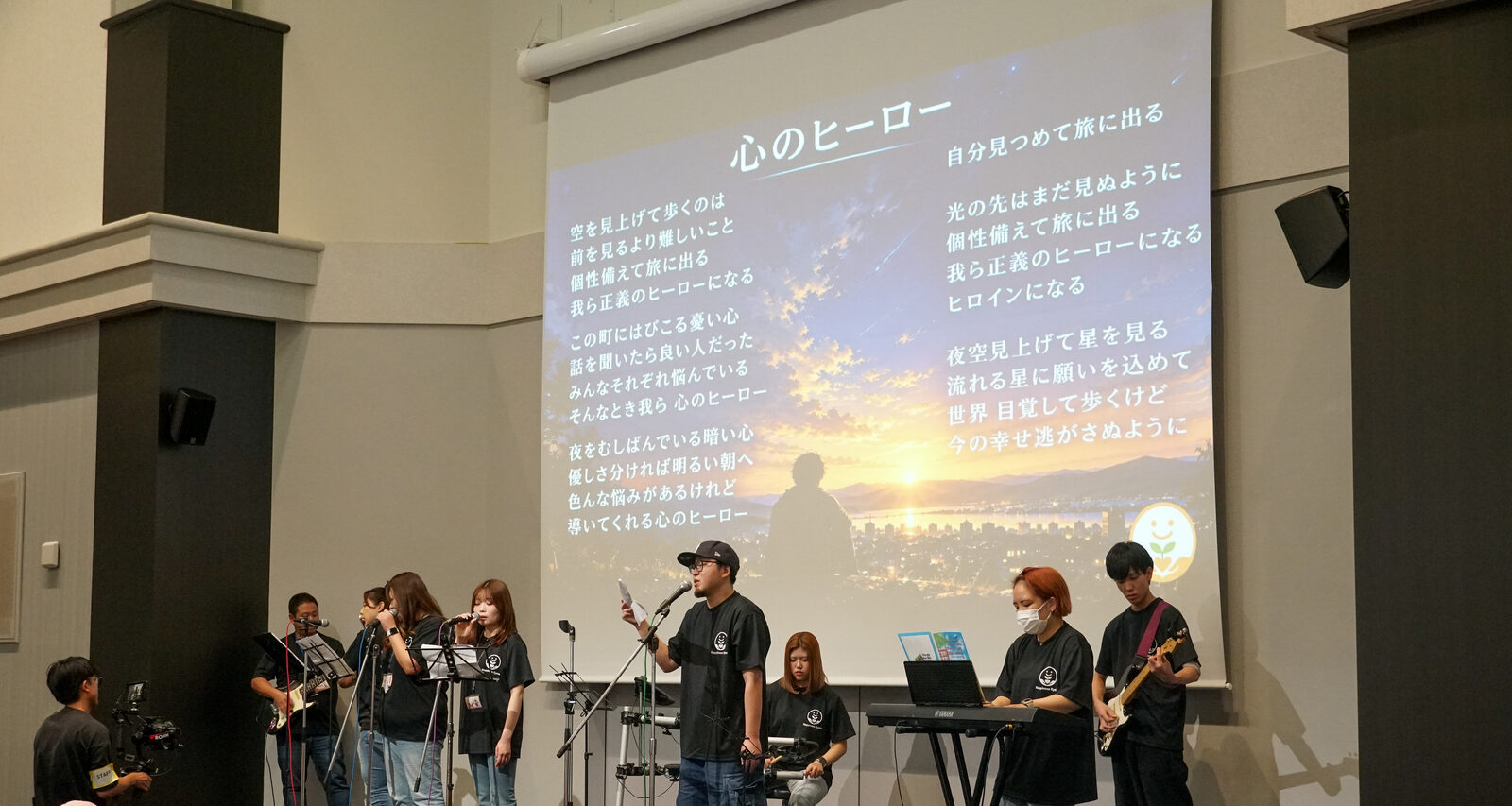 バンドが演奏する背後のスクリーンに、夕景の写真と「心のヒーロー」の歌詞が映し出されている。