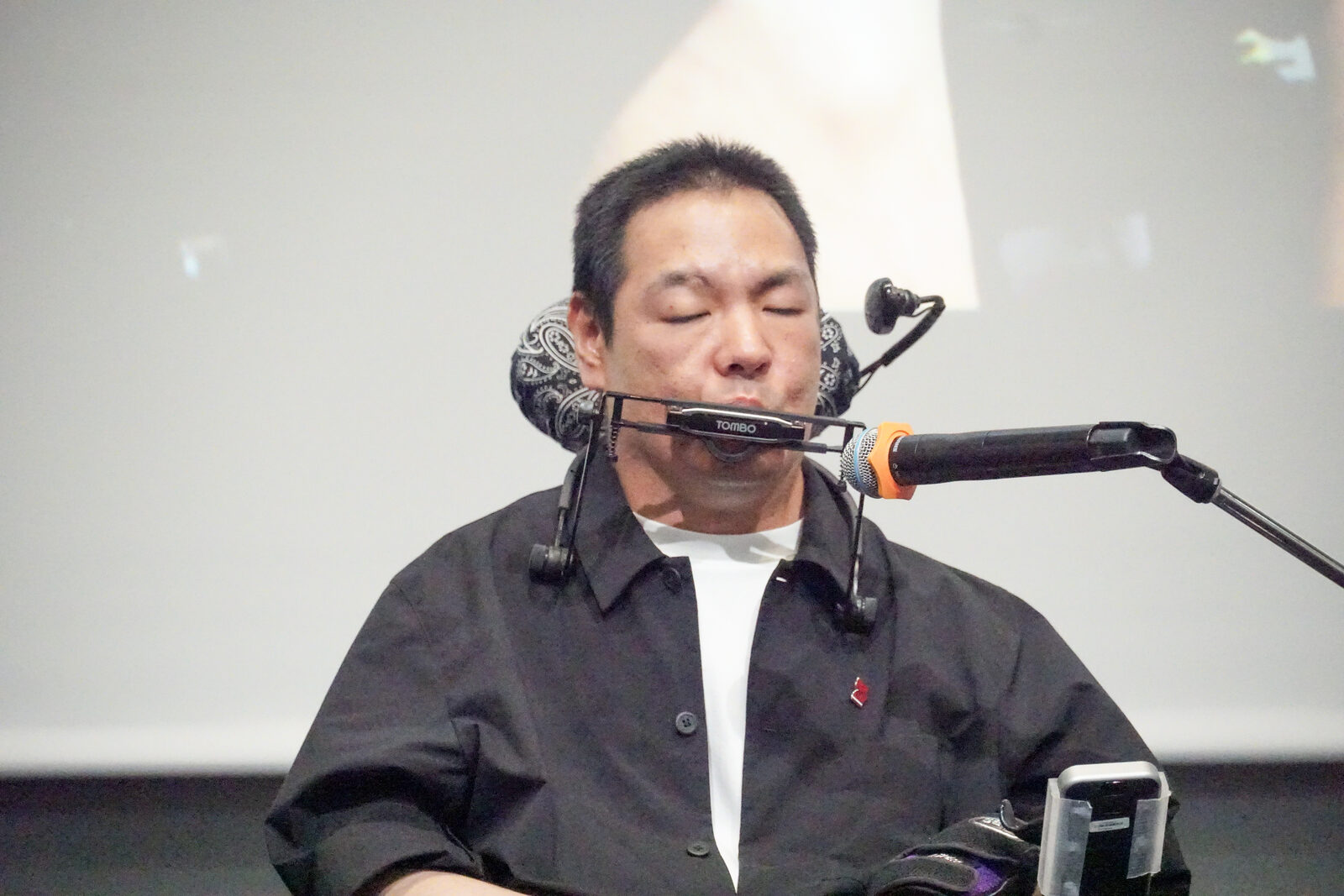 伊藤靖幸さんが口元に固定したハーモニカを演奏している。目を閉じた横顔。