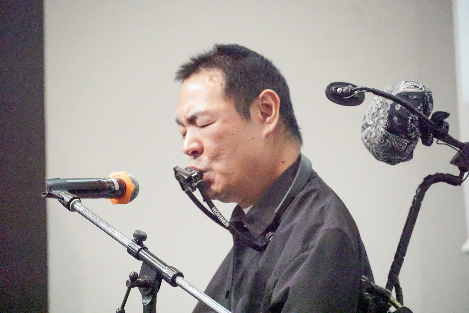 伊藤靖幸さんがマイクに向かってハーモニカを吹いている、別アングルの写真。